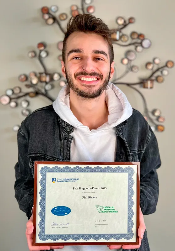 Phil Rivière tient dans ses mains le Prix Huguette-Parent 2023, prix créé par le Réseau du Patrimoine franco-ontarien en collaboration avec l’Université Laurentienne.