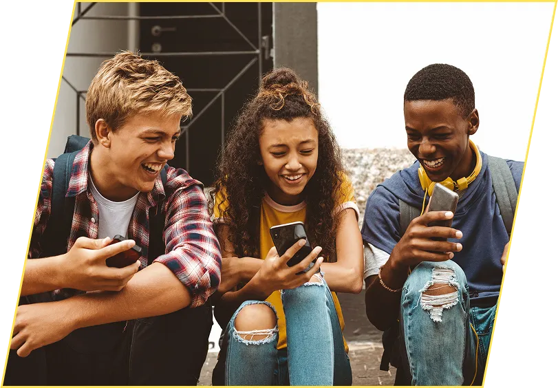 Trois jeunes sont assis à l’extérieur. Ils consultent leur téléphone cellulaire. Ils rient.