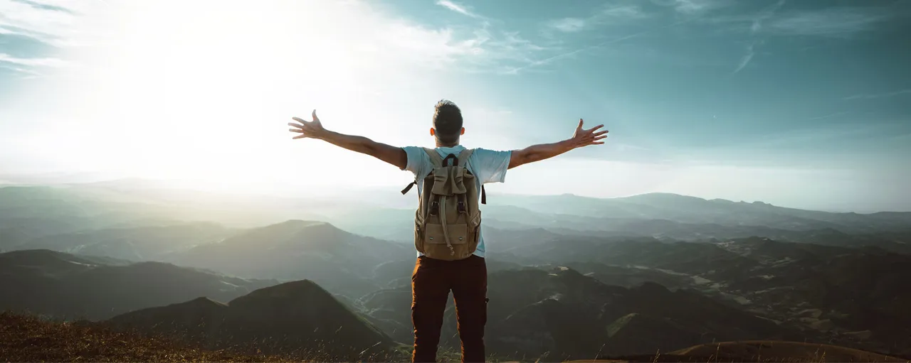 Un homme porte un sac à dos. Il a les bras étendus de chaque côté de son corps. Il est au sommet d’une montagne.