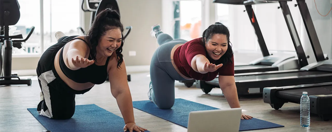 Deux femmes font une pose de yoga en regardant l’écran d’un ordinateur portable. Elles sourient.