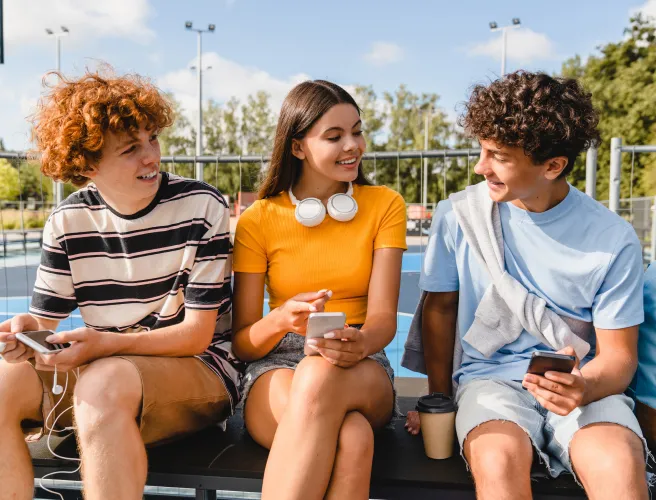 Une adolescente et deux adolescents sont assis sur un banc à l’extérieur. Ils tiennent dans leurs mains un cellulaire. Ils parlent ensemble.