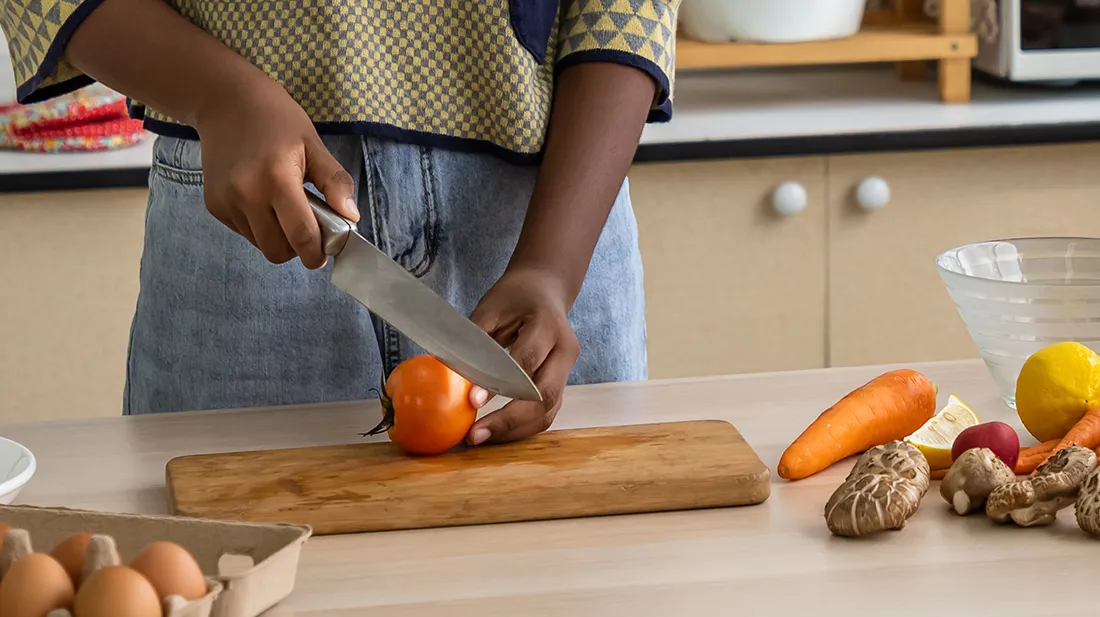 Une personne coupe une tomate en utilisant un gros couteau et une planche de bois. Il y a d’autres légumes près de la planche.