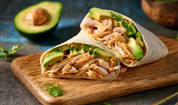 Deux demi-sandwichs faits de poulet et d’avocats enroulés dans une tortilla, présentés sur une planche de bois.