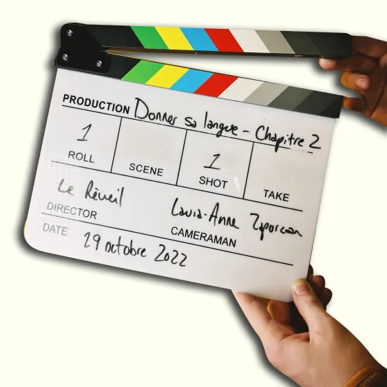 Une claquette affichant : « Production : Donner sa langue – Chapitre 2. Roll 1, Séquence 1, Prise 1. Directeur : Le réveil. Caméra : Laura-Anne Zaporzan. Date : 29 oct. 2022 ».