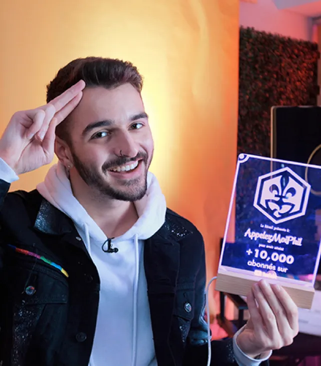Phil Rivière salue en tenant un trophée YouTube. Celui-ci affiche un logo (fleur de lys et trille blanc dans un hexagone), le nom de la chaîne Appelez-moi Phil et la mention « Plus de 10 000 abonnés sur YouTube ».